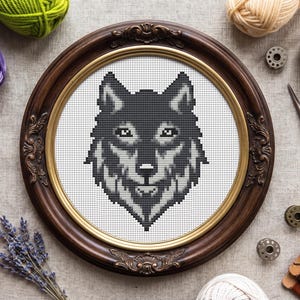 May include: A framed cross-stitch of a wolf's head in shades of gray and black. The artwork is displayed in a dark brown, ornate oval frame with a gold inner border. The background includes yarn, scissors, and other craft supplies.