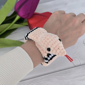 May include: A pink crochet pattern snake slap bracelet with black and white checkered accents. The snake has a red tongue and a black eye.