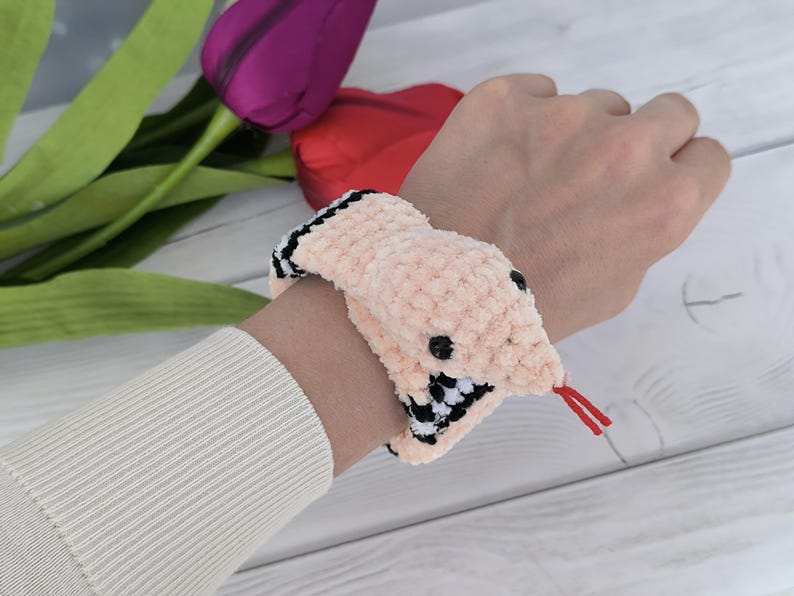 May include: A pink crochet pattern snake slap bracelet with black and white checkered accents. The snake has a red tongue and a black eye.