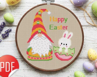 May include: A cross-stitch pattern in a wooden embroidery hoop featuring a gnome with a tall striped hat and a white beard next to a small white bunny sitting in a pink and green egg. The text Happy Easter is stitched in colorful letters.