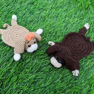 "A top-down view of two crocheted coasters shaped like dogs laying on a green grass surface. On the left is a tan crocheted dog coaster wearing an orange bow. On the right is a dark brown crocheted dog coaster. Both coasters feature small crocheted bodies, floppy ears, white paws, small safety eyes, and little white muzzles. These handmade items are based on a crochet pattern for dog coasters."