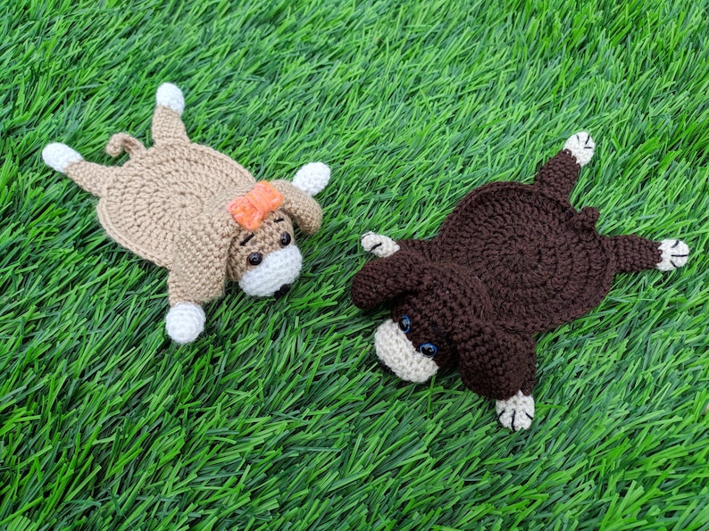 "A top-down view of two crocheted coasters shaped like dogs laying on a green grass surface. On the left is a tan crocheted dog coaster wearing an orange bow. On the right is a dark brown crocheted dog coaster. Both coasters feature small crocheted bodies, floppy ears, white paws, small safety eyes, and little white muzzles. These handmade items are based on a crochet pattern for dog coasters."
