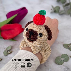 May include: A crocheted pug dog wristband in tan and brown, with a red and green pom-pom on its head. The wristband is soft and plush, with black eyes and a dark brown nose. The text "Crochet Pattern" is visible at the bottom.