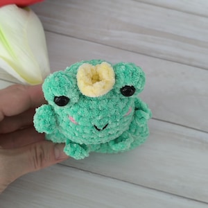 A plush turquoise amigurumi frog with a small crown, made using a slap bracelet crochet pattern, worn on a wrist. This Frog slap bracelet pattern is available in English.