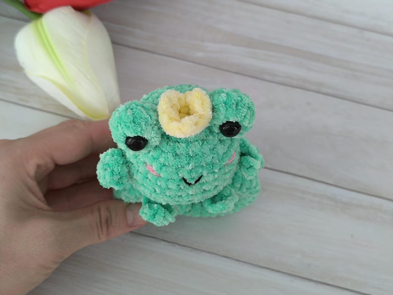 A plush turquoise amigurumi frog with a small crown, made using a slap bracelet crochet pattern, worn on a wrist. This Frog slap bracelet pattern is available in English.