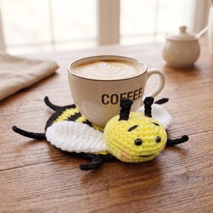 May include: A beige coffee mug with the word "COFFEE" printed on the side, resting on a handmade crochet bee coaster. The coaster features yellow, black, and white yarn. The mug contains a latte with a leaf design.