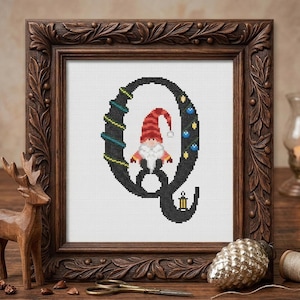 May include: A cross-stitch embroidery of the letter Q in black, decorated with green garland, blue ornaments, and a small lantern. A gnome with a red striped hat and white beard is positioned in the center of the letter, all framed in dark wood.