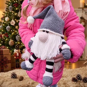 A crochet pattern gnome doll with a gray hat and body, white beard, and pink nose. The gnome has black and white striped legs and is wearing gray shoes.