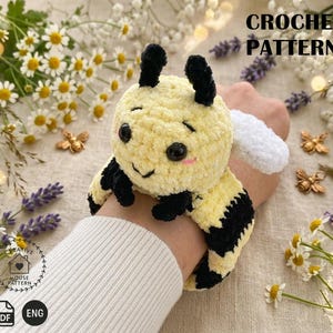 May include: A crocheted bee toy with yellow and black stripes, black antennae, and a white wing. The bee has a smiling face with pink cheeks. The image also includes the text "CROCHET PATTERN".