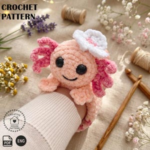 May include: A crocheted axolotl plushie in soft peach and pink hues, with a white flower accent. The axolotl has black eyes and a smiling mouth. The image includes the text "CROCHET PATTERN" and a logo with "CREATIVE HOUSE PATTERN".