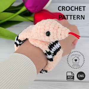 May include: A crochet pattern for a plush snake toy. The snake is made of soft peach-colored yarn with black and white striped sections, featuring a small black eye and a red yarn tongue. The text CROCHET PATTERN, CREATIVE HOUSE PATTERN, PDF, and ENG are visible.
