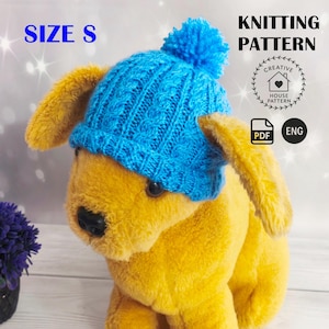 Könnte beinhalten: Eine blaue Strickmütze für Hunde mit Bommel, beschriftet mit "SIZE S". Die Mütze ist Teil eines Strickmusters, wie durch den Text "KNITTING PATTERN" und ein "PDF ENG"-Symbol angezeigt wird. Die Mütze wird von einem gelben Stofftier getragen.