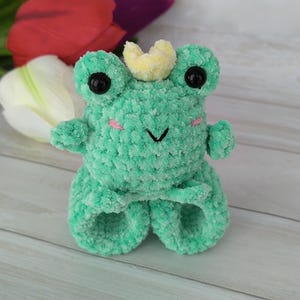 A plush turquoise amigurumi frog with a small crown, made using a slap bracelet crochet pattern, worn on a wrist. Below are colorful tulips. This Frog slap bracelet pattern is available in English.