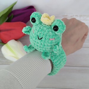 From "Creative House Pattern," an ENG PDF download for this charming plush frog slap bracelet. The crocheted frog, featuring colorful yarn and cute details, is shown on a modeled wrist. Tulips fill the background. Perfect for learning to make your own reusable accessories using the "slap bracelet crochet pattern Frog.