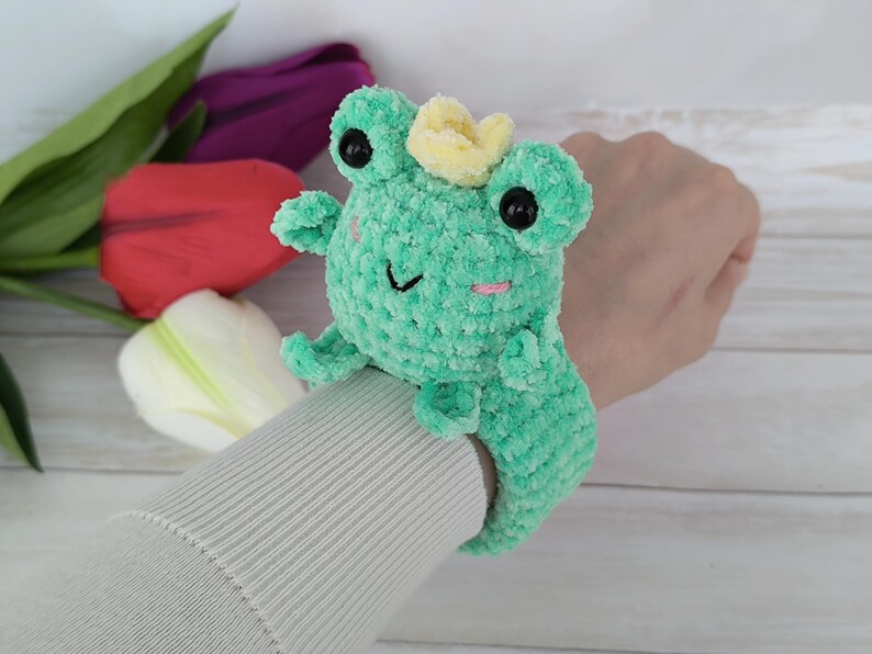 From "Creative House Pattern," an ENG PDF download for this charming plush frog slap bracelet. The crocheted frog, featuring colorful yarn and cute details, is shown on a modeled wrist. Tulips fill the background. Perfect for learning to make your own reusable accessories using the "slap bracelet crochet pattern Frog.