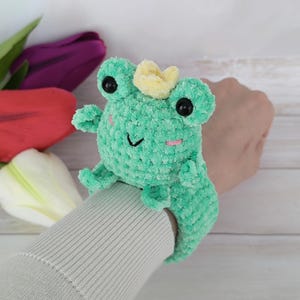 A photo showcasing a completed project from the "Frog slap bracelet crochet pattern." A woman's wrist models a cute, soft turquoise crocheted frog with a small yellow crown, safety eyes, and little pink cheek details. The plush frog is securely fastened around the wrist like a slap bracelet. The background features white and pink tulips.