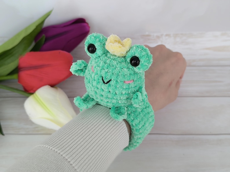 A photo showcasing a completed project from the "Frog slap bracelet crochet pattern." A woman's wrist models a cute, soft turquoise crocheted frog with a small yellow crown, safety eyes, and little pink cheek details. The plush frog is securely fastened around the wrist like a slap bracelet. The background features white and pink tulips.