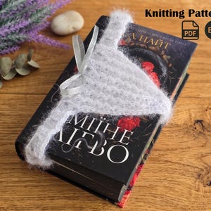 Puede incluir: Un marcapáginas gris tejido descansa sobre un libro con una cubierta negra. El libro tiene detalles florales rojos y texto en un idioma extranjero. El marcapáginas está atado con una cinta plateada. La imagen también incluye el texto "Knitting Pattern" y "PDF ENG".