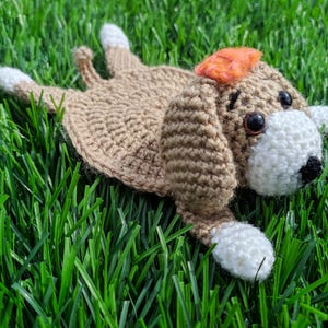 A handmade crochet dog coaster in a lying-down pose. The coaster is primarily tan with white paws and a white face. It has a small orange bow and black eyes.