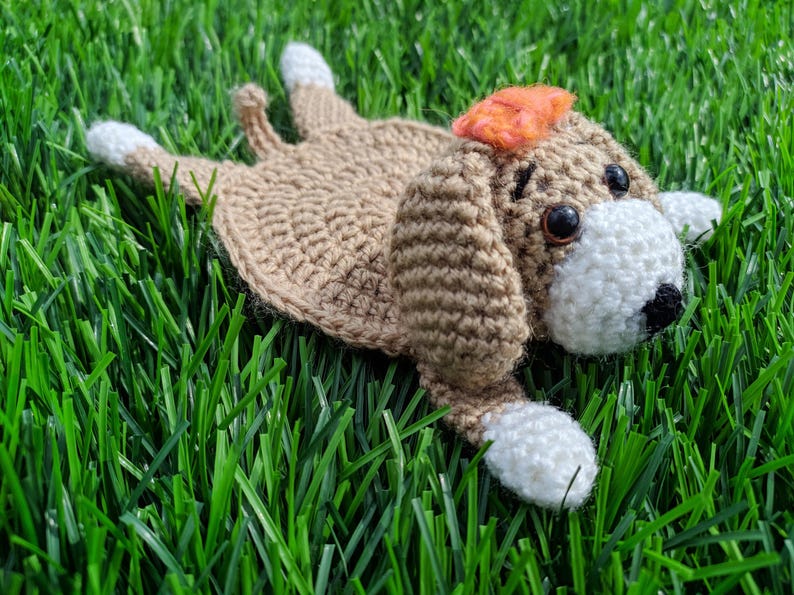 A handmade crochet dog coaster in a lying-down pose. The coaster is primarily tan with white paws and a white face. It has a small orange bow and black eyes.