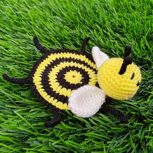 May include: A handmade crochet bee mug rug with a yellow head and body, black and yellow striped body, white wings, and black antennae. The bee is lying on a bed of green artificial grass.
