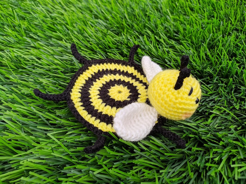 May include: A handmade crochet bee mug rug with a yellow head and body, black and yellow striped body, white wings, and black antennae. The bee is lying on a bed of green artificial grass.