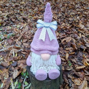 May include: A handmade crochet gnome doll with a lavender hat and matching feet. The gnome has a white beard, a peach-colored nose, and a silver bow. The doll is sitting on a tree stump surrounded by fallen leaves.