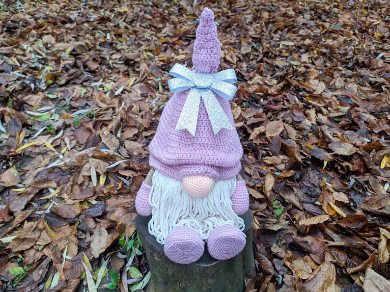 May include: A handmade crochet gnome doll with a lavender hat and matching feet. The gnome has a white beard, a peach-colored nose, and a silver bow. The doll is sitting on a tree stump surrounded by fallen leaves.
