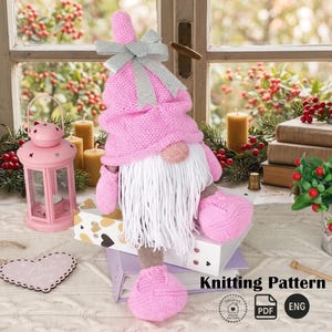 Könnte beinhalten: Eine rosa gestrickte Zwergpuppe mit weißem Bart und einer silbernen Schleife auf dem Hut. Der Zwerg sitzt auf einem Stapel Bücher und einer dekorativen Schachtel. Das Bild enthält auch eine rosa Laterne und Kerzen.