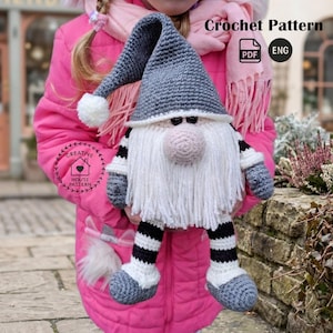 Könnte beinhalten: Eine handgefertigte gehäkelte Zwergenpuppe mit grauem Hut, weißem Bart und gestreiften Beinen. Die Puppe hat eine rosa Nase und hält ein Crochet Pattern PDF. Die Puppe ist etwa 30 cm groß.