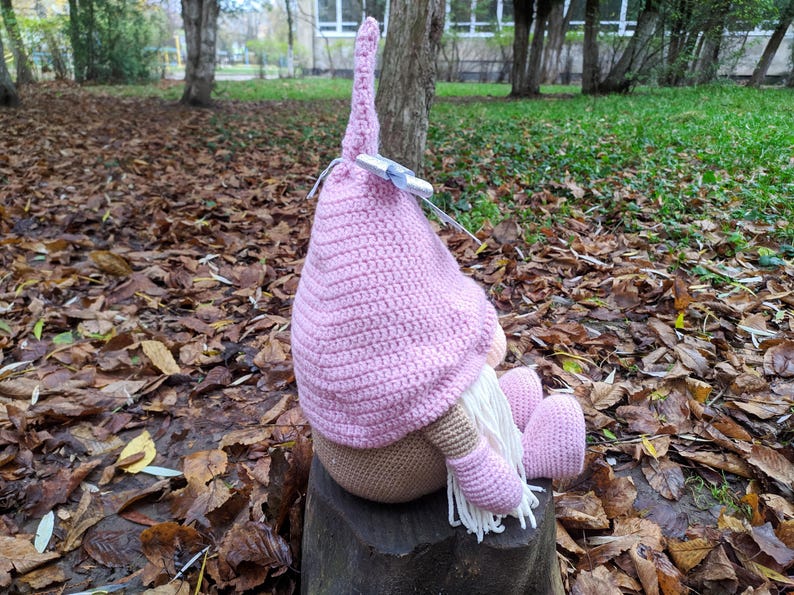 May include: A handmade crochet gnome doll with a pink hat and matching feet and arms. The gnome has a beige body and a long white beard. It is sitting on a tree stump outdoors, surrounded by fallen leaves.