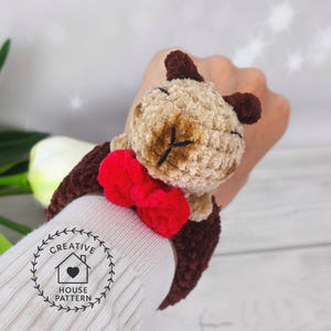May include: Sensory Capybara Fidget Toy Crochet Pattern. The Capybara is attached to a brown wristband. The wristband is shown on a person's wrist.