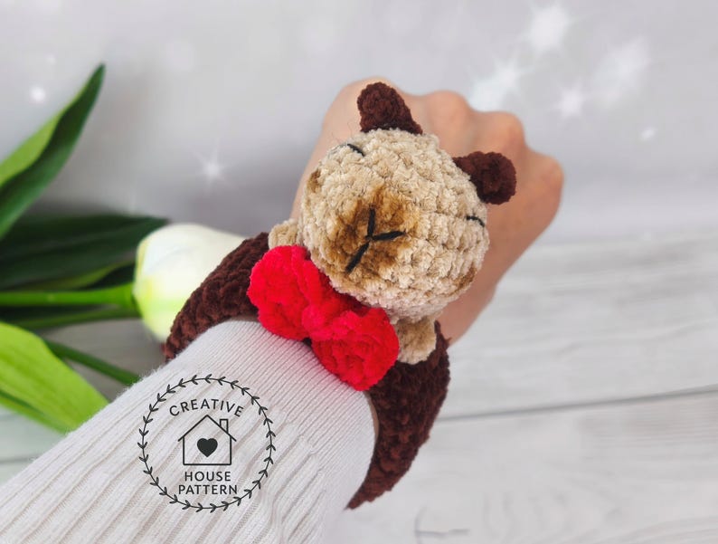 May include: Sensory Capybara Fidget Toy Crochet Pattern. The Capybara is attached to a brown wristband. The wristband is shown on a person's wrist.