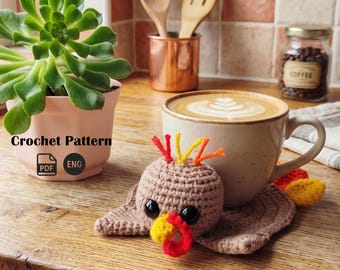 May include: A crocheted turkey coaster sits next to a coffee mug with latte art. The turkey has a brown body, a yellow and red beak, and colorful feather accents. A small succulent plant in a pink pot is in the background. The text "Crochet Pattern" is visible.