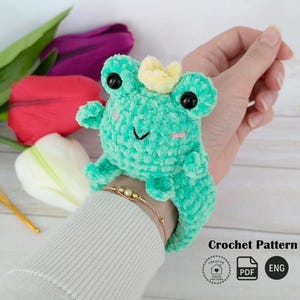 A plush turquoise amigurumi frog with a small crown, made using a slap bracelet crochet pattern, worn on a wrist. Below are colorful tulips and the text "Crochet Pattern" with "PDF" and "ENG" icons. This Frog slap bracelet pattern is available in English.