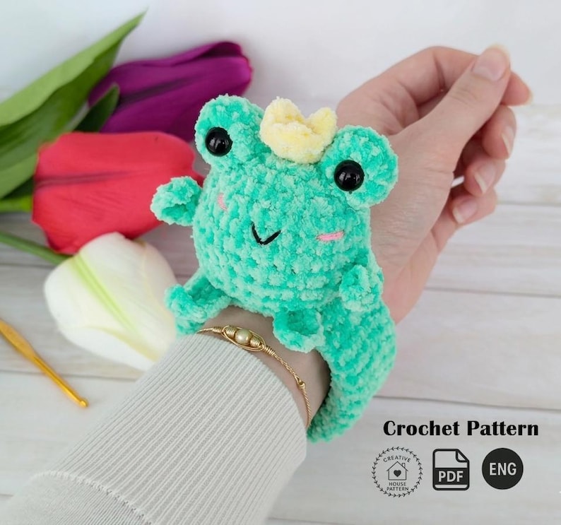 A plush turquoise amigurumi frog with a small crown, made using a slap bracelet crochet pattern, worn on a wrist. Below are colorful tulips and the text "Crochet Pattern" with "PDF" and "ENG" icons. This Frog slap bracelet pattern is available in English.
