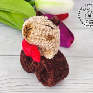 Sensory Capybara Fidget Toy Crochet Pattern with a red bow tie. The  Capybara has closed eyes and is attached to a dark brown wristband. The image also includes the text "Crochet Pattern". The Capybara is made of soft, textured fabric. The toy is sitting on a white wooden surface.