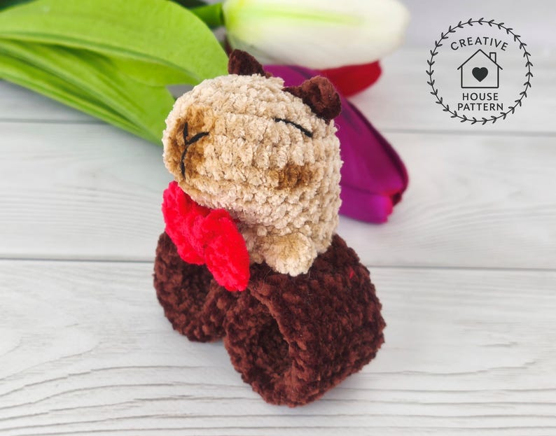 Sensory Capybara Fidget Toy Crochet Pattern with a red bow tie. The  Capybara has closed eyes and is attached to a dark brown wristband. The image also includes the text "Crochet Pattern". The Capybara is made of soft, textured fabric. The toy is sitting on a white wooden surface.