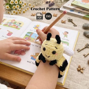 May include: A crocheted bee wristband in yellow and black, with white wings, worn on a wrist. The bee has a smiling face. The image also shows a pencil, an open book, and the text "Crochet Pattern".