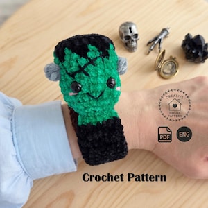 May include: A green and black crochet monster wrist cuff featuring a stitched forehead detail, black safety eyes, and small grey side bolts. The design includes a Creative House Pattern logo, a PDF icon, an ENG label, and the text Crochet Pattern.