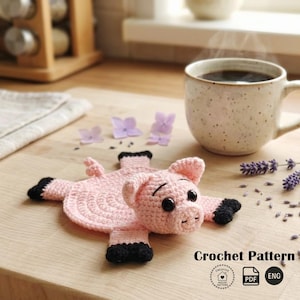 May include: A handmade pink crochet pig coaster with black hooves and ears. The pig has black button eyes and a small snout. A cup of coffee sits next to the coaster. The words "Crochet Pattern" are at the bottom of the image.