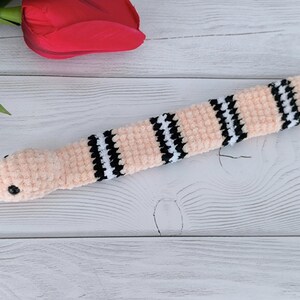 May include: A pink crochet pattern snake slap bracelet with black and white checkered accents. The snake has a red tongue and a black eye.
