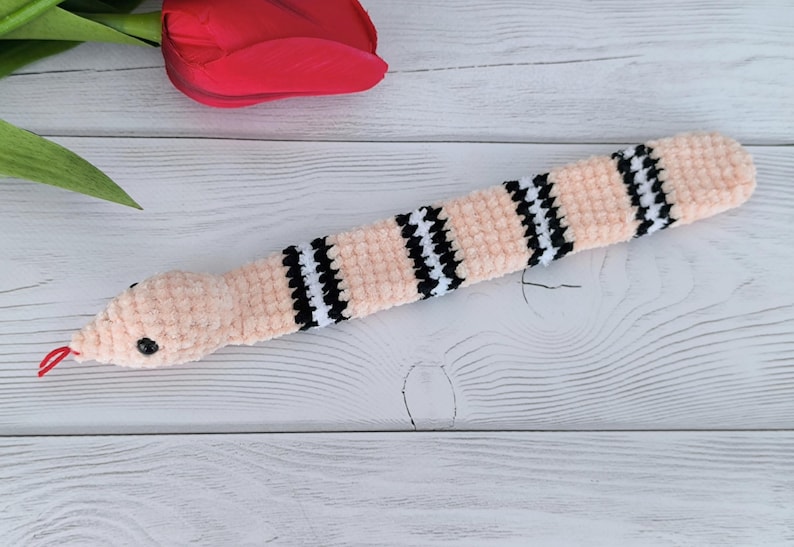 May include: A pink crochet pattern snake slap bracelet with black and white checkered accents. The snake has a red tongue and a black eye.