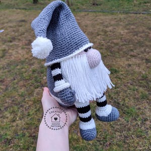May include: A handmade crochet gnome doll with a gray hat and body, white beard, and pink nose. The gnome has black and white striped legs and is wearing gray shoes. The doll is a decorative item.
