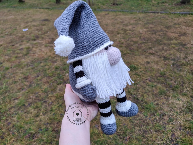May include: A handmade crochet gnome doll with a gray hat and body, white beard, and pink nose. The gnome has black and white striped legs and is wearing gray shoes. The doll is a decorative item.