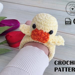 May include: A crocheted yellow duck with an orange beak and black eyes. The duck is holding a person's arm. The image also includes the text "CROCHET PATTERN" and a logo with the words "CREATIVE HOUSE PATTERN".