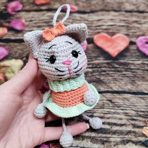 Peut inclure: Une poup&eacute;e chat au crochet faite &agrave; la main, avec un corps gris, un nez rose et des yeux noirs. Elle porte une robe verte et orange et un petit n&oelig;ud orange. La poup&eacute;e est suspendue par une ficelle blanche.