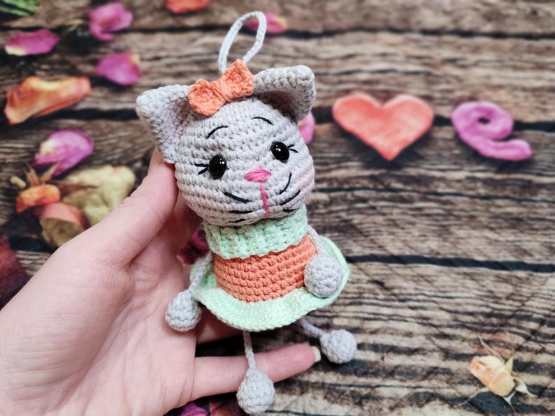 Peut inclure: Une poup&eacute;e chat au crochet faite &agrave; la main, avec un corps gris, un nez rose et des yeux noirs. Elle porte une robe verte et orange et un petit n&oelig;ud orange. La poup&eacute;e est suspendue par une ficelle blanche.
