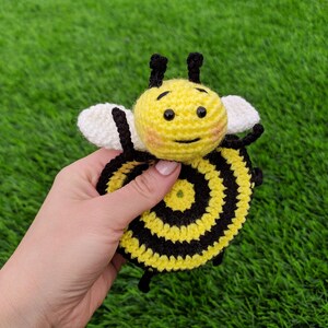 May include: A handmade crochet bee coaster with a yellow and black striped body, white wings, and black antennae. The bee has a smiling face with rosy cheeks. The toy is held against a backdrop of green grass.