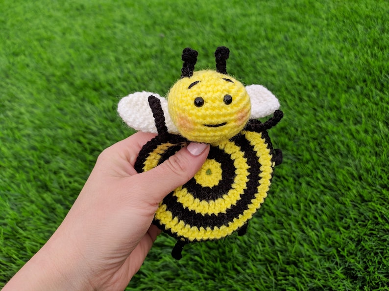 May include: A handmade crochet bee coaster with a yellow and black striped body, white wings, and black antennae. The bee has a smiling face with rosy cheeks. The toy is held against a backdrop of green grass.